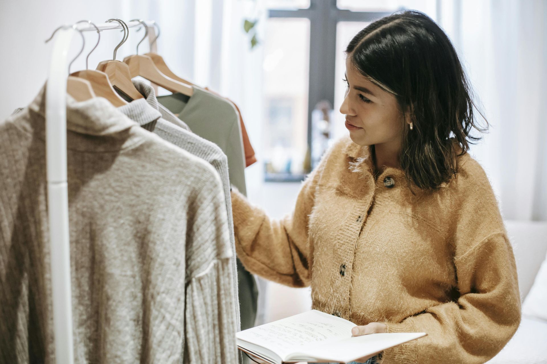 A woman designer examines clothing on a rack indoors, holding a notebook.